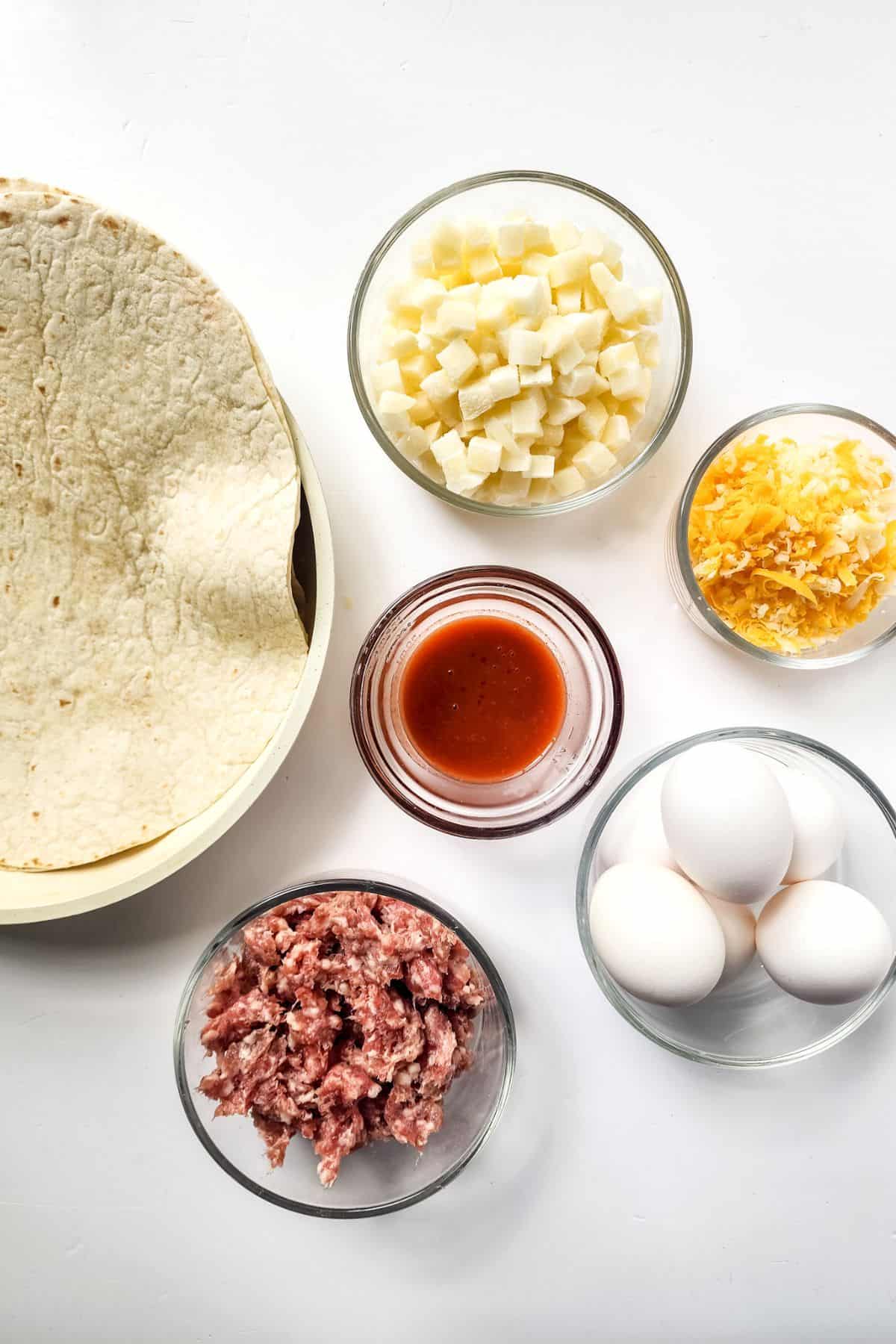 A tortilla with bowls of cubed cheese, shredded cheese, tomato sauce, raw ground meat, and whole eggs on a white surface—perfect ingredients for assembling your own Breakfast Crunchwrap.