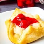 A cherry cheese danish pastry with a golden crust and cherry topping is served on a white plate, reminiscent of a Cheesecake Bites Recipe, with a jar of cherries in the background.