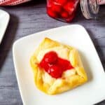 A square white plate holds a cheesecake bites recipe pastry, topped with cream and two cherries; in the background, a jar of cherry topping and a red-and-white cloth are visible.