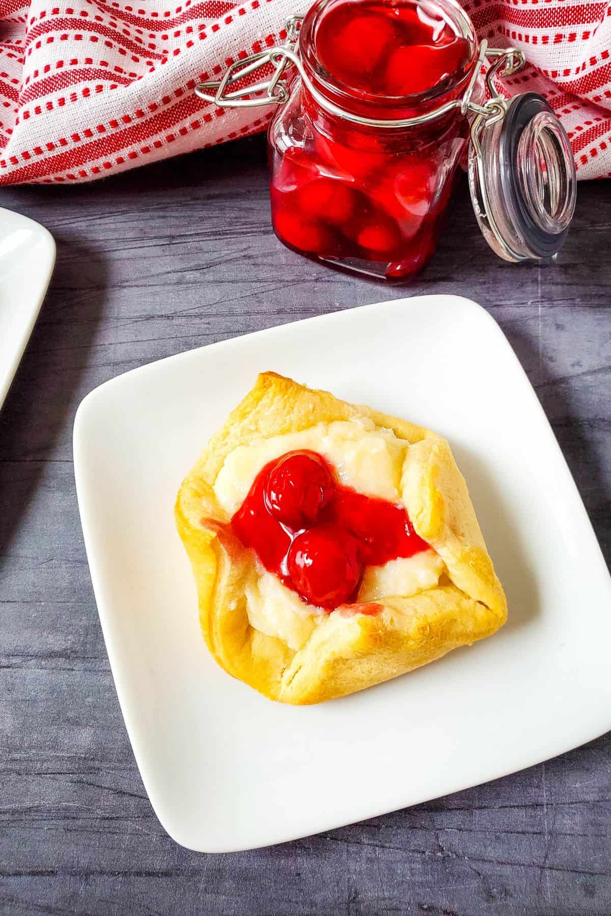 A square white plate holds a cheesecake bites recipe pastry, topped with cream and two cherries; in the background, a jar of cherry topping and a red-and-white cloth are visible.