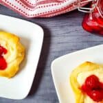 Two treats on white plates beside a red-and-white cloth and a jar of cherry filling on a gray surface, perfect for anyone who loves a delicious Cheesecake Bites Recipe.