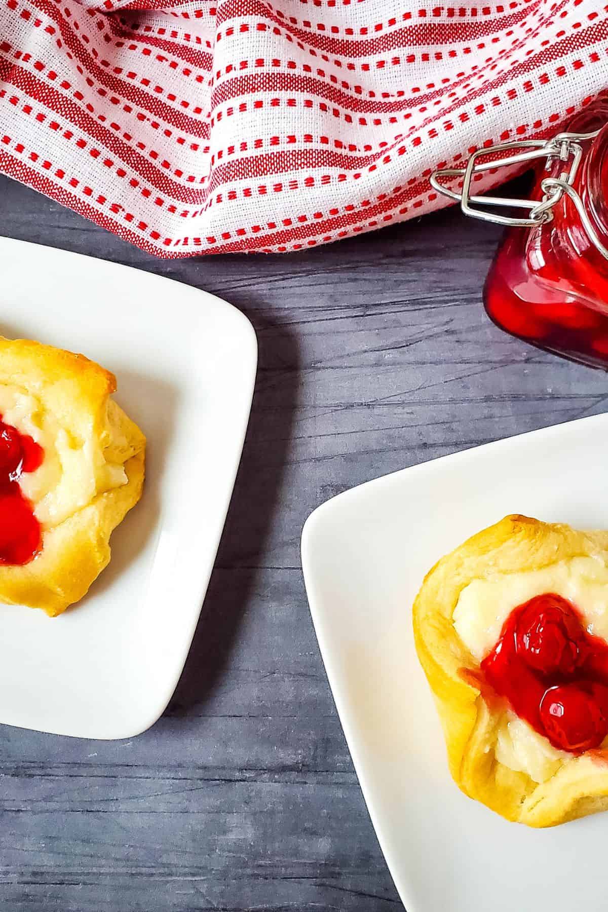 Two treats on white plates beside a red-and-white cloth and a jar of cherry filling on a gray surface, perfect for anyone who loves a delicious Cheesecake Bites Recipe.