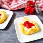 A square cream cheese danish topped with cherry filling on a white plate, reminiscent of a Cheesecake Bites Recipe, with a jar of cherry preserves and a red-striped cloth in the background.