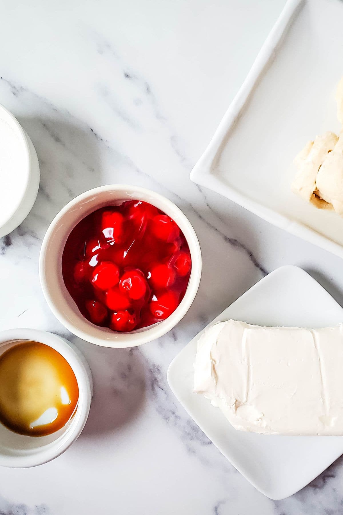 A bowl of cherry pie filling, a block of cream cheese on a plate, vanilla extract in a small dish, and biscuit dough on a white tray set the scene for this delicious Cheesecake Bites Recipe, all arranged on a marble surface.