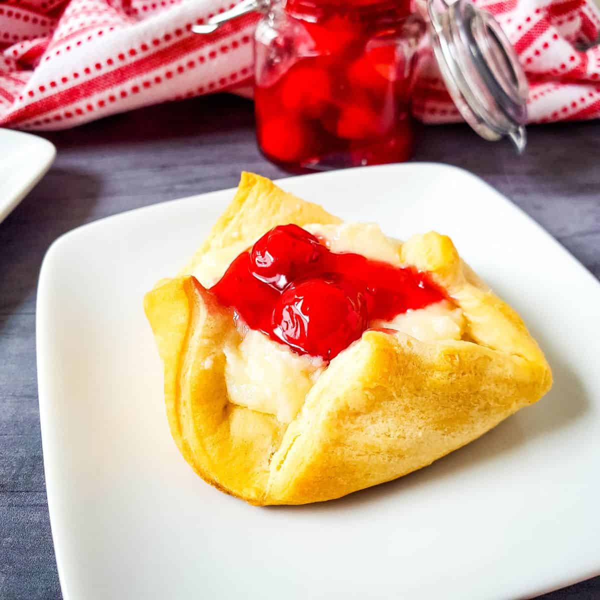A pastry topped with cream filling and cherry pie filling sits on a white plate, showcasing a delicious Cheesecake Bites Recipe, with a jar of cherries and a red-striped cloth in the background.