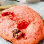 A close-up of a Cherry Chip Cookie with visible chocolate chips and red fruit pieces, placed on parchment paper. A cloth is partially visible in the background.