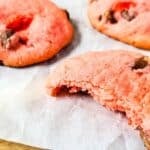 Three cherry chip cookies with chocolate chips rest on parchment paper, one with a bite taken out of it.