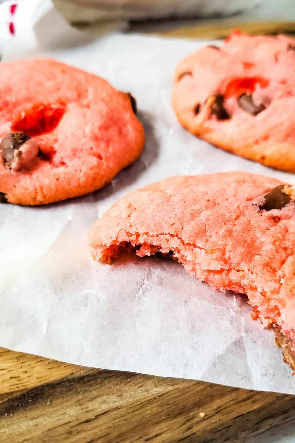 Three cherry chip cookies with chocolate chips rest on parchment paper, one with a bite taken out of it.