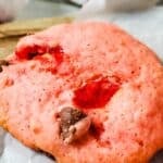 A close-up of a Cherry Chip Cookie with chocolate chips and visible pieces of cherry, resting on parchment paper.