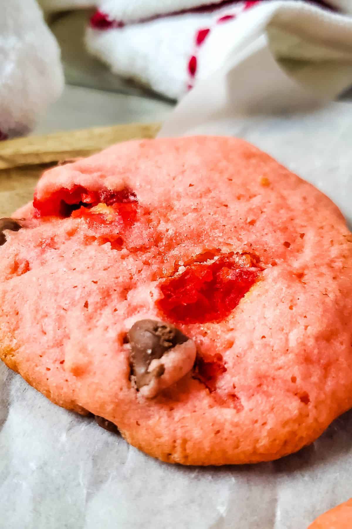 A close-up of a Cherry Chip Cookie with chocolate chips and visible pieces of cherry, resting on parchment paper.