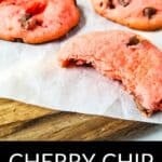 Three pink Cherry Chip Cookies, one with a bite taken out, rest on parchment paper atop a wooden surface.