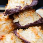 A close-up of coconut and almond macaroons stacked in a pile, each with toasted coconut on top and the bottoms dipped in chocolate.