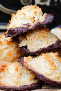 A close-up of coconut and almond macaroons stacked in a pile, each with toasted coconut on top and the bottoms dipped in chocolate.