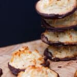 A stack of coconut and almond macaroons dipped in chocolate sits on a wooden surface, with two macaroons lying in front.