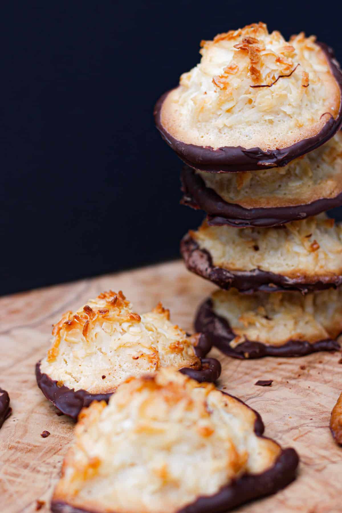 A stack of coconut and almond macaroons dipped in chocolate sits on a wooden surface, with two macaroons lying in front.