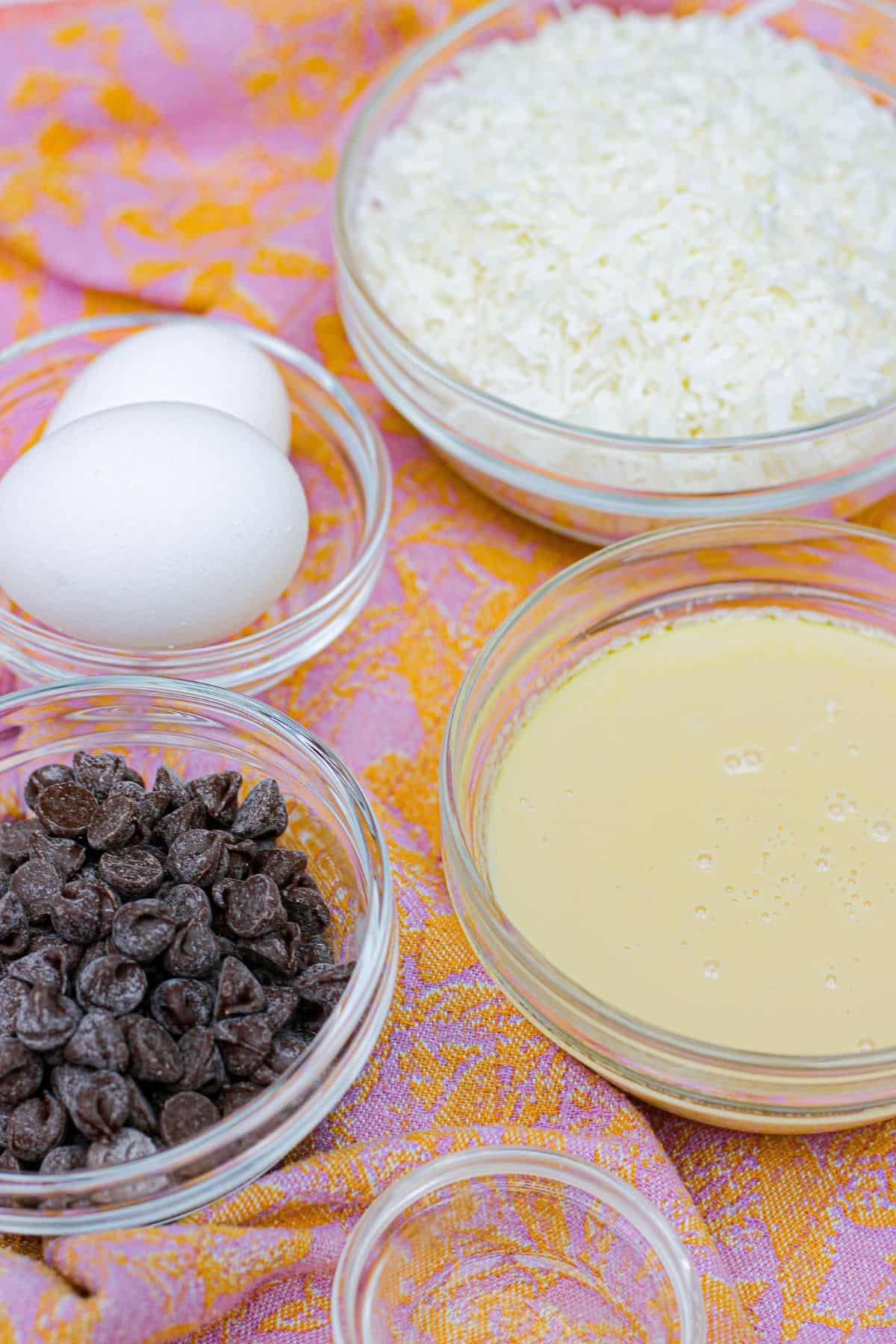 Bowls of shredded coconut, chocolate chips, sweetened condensed milk, and two whole eggs are displayed on a pink and orange patterned surface, ready for baking .
