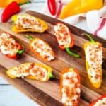 Stuffed Mini Peppers halved and filled with a creamy mixture, topped with crispy bacon bits, are artfully arranged on a wooden serving board with whole peppers and a towel in the background.