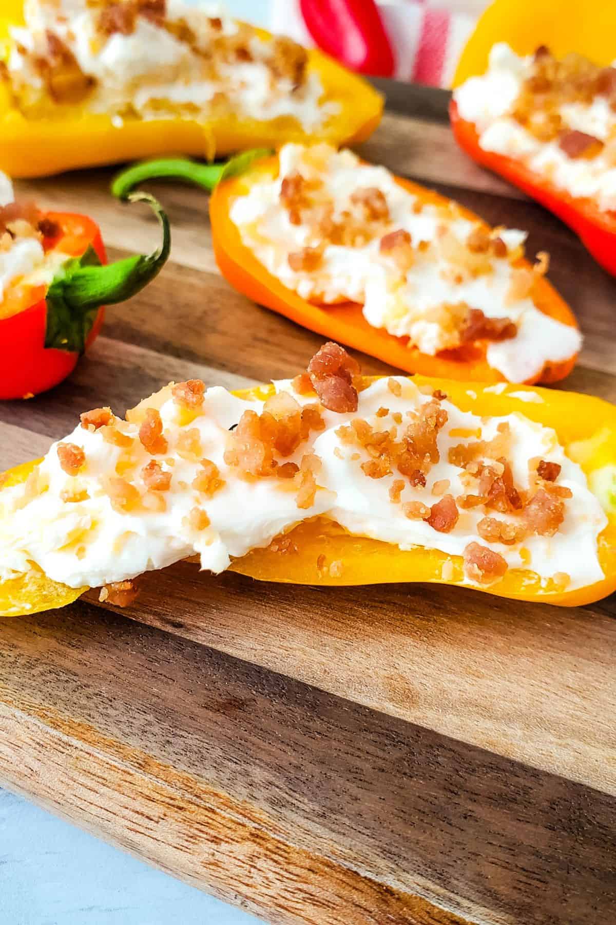 Stuffed Mini Peppers are halved and filled with a creamy mixture, then topped with bacon bits and beautifully arranged on a wooden serving board.