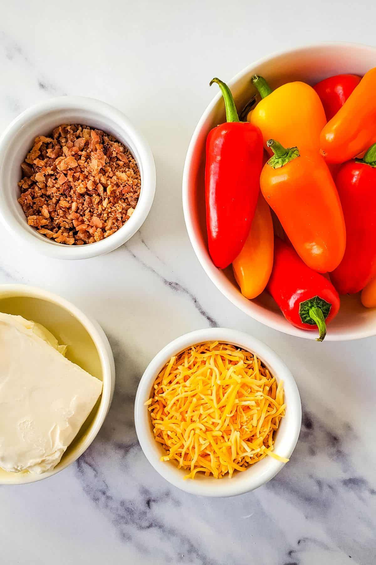 Four bowls on a marble surface hold the key ingredients crumbled bacon, mini bell peppers, cream cheese, and shredded cheddar cheese.