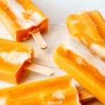 Five orange creamsicle popsicles are arranged on a light-colored surface. Some popsicles are partially eaten, revealing their creamy texture.