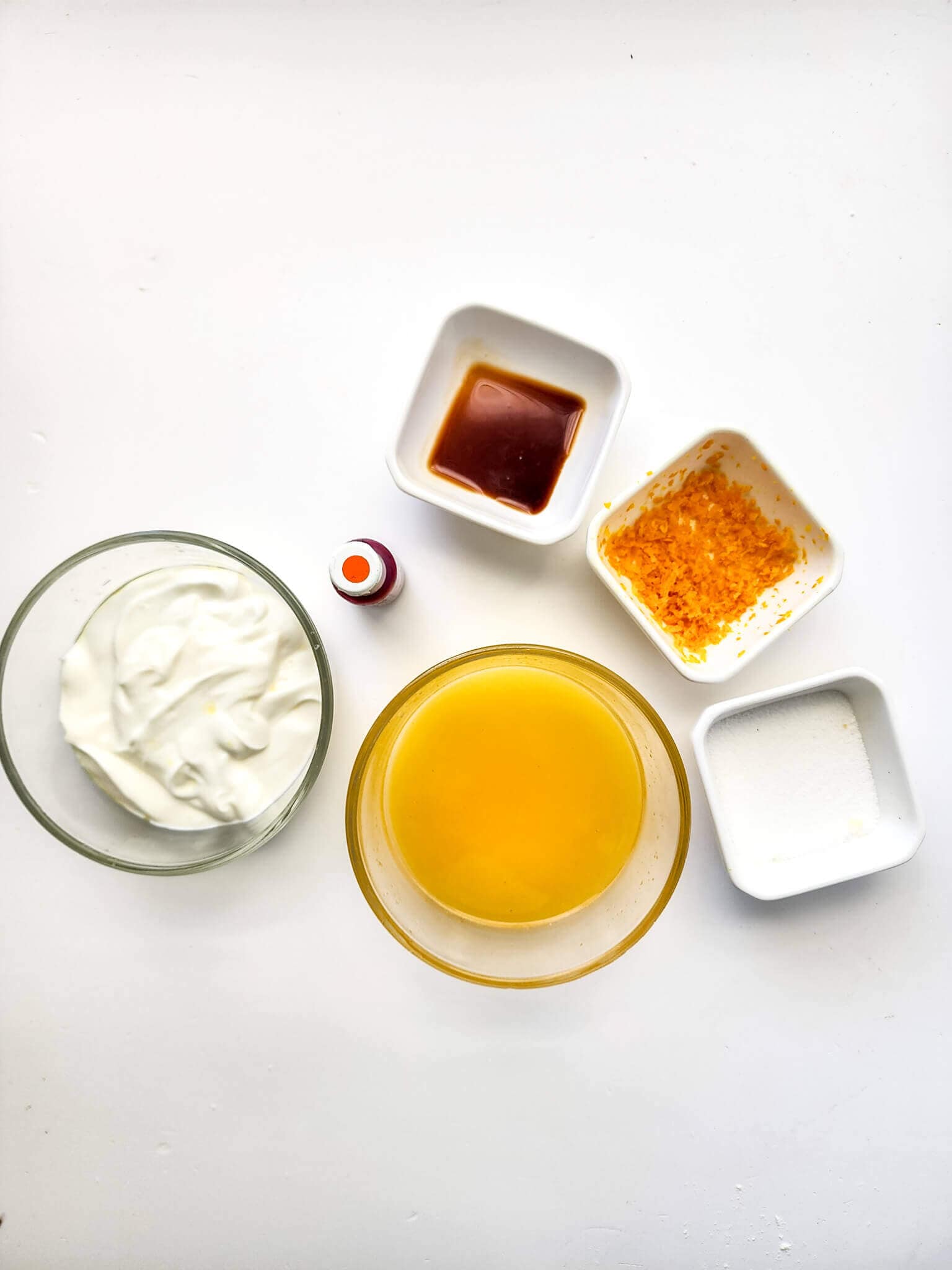 Ingredients for Orange Popsicles include a small bowl of yogurt, a bowl of orange juice, a small bottle of red liquid, a dish of vanilla extract, a dish of orange zest, and a dish of sugar.
