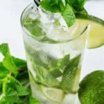 A glass of Cuban Mojito with ice, mint leaves, a lime wedge, and a black and white striped straw. A whole lime and mint sprigs are placed next to the glass on a white surface.