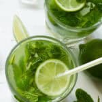 Two glasses of a refreshing Cuban Mojito with lime slices, mint leaves, and ice cubes on a white surface, accompanied by a wedge of lime and an ice cube.