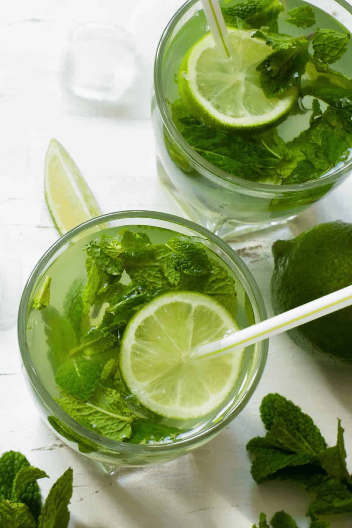 Two glasses of a refreshing Cuban Mojito with lime slices, mint leaves, and ice cubes on a white surface, accompanied by a wedge of lime and an ice cube.
