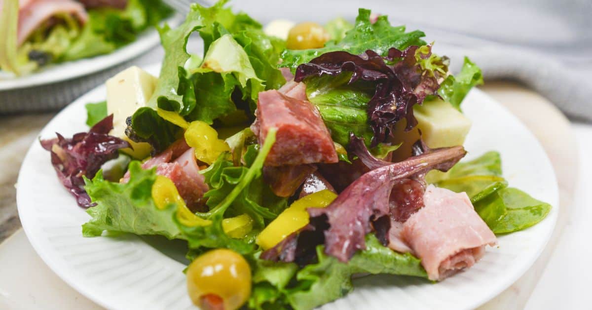 A plate of Antipasto Misto features mixed leafy greens salad topped with cubes of cheese, rolled slices of ham, salami, green olives, and yellow pepper strips.