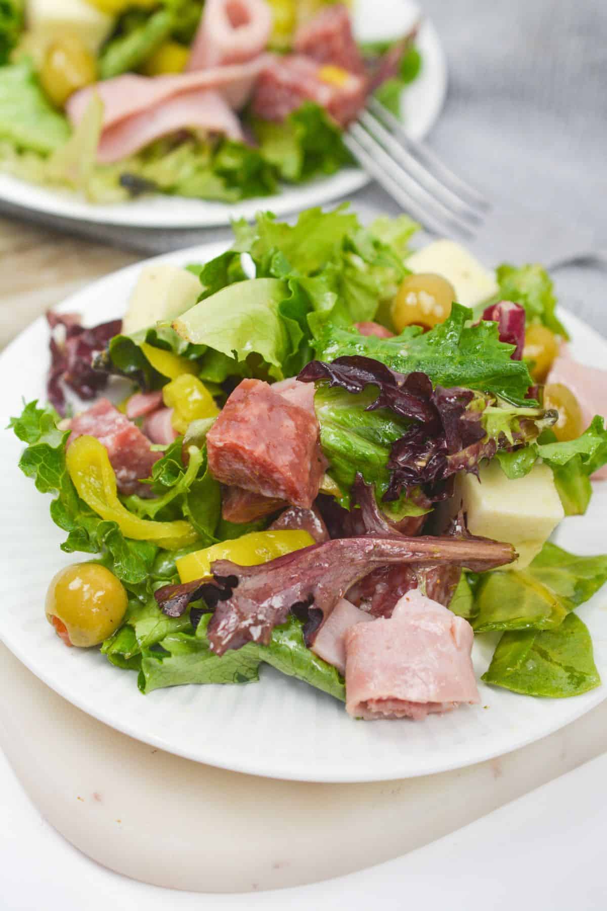 A plate of Antipasto Misto featuring mixed green salad with sliced deli meats, green olives, cheese cubes, and yellow peppers.