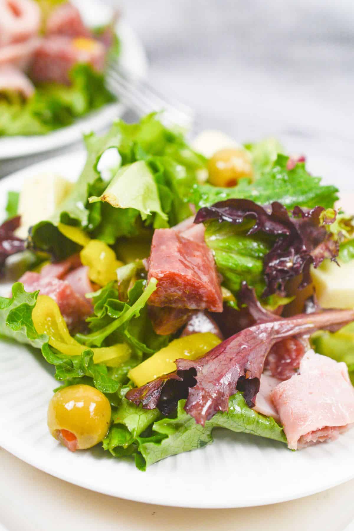 A close-up of an Antipasto Misto salad with mixed greens, sliced meats, green olives, yellow peppers, and cubes of cheese on a white plate.