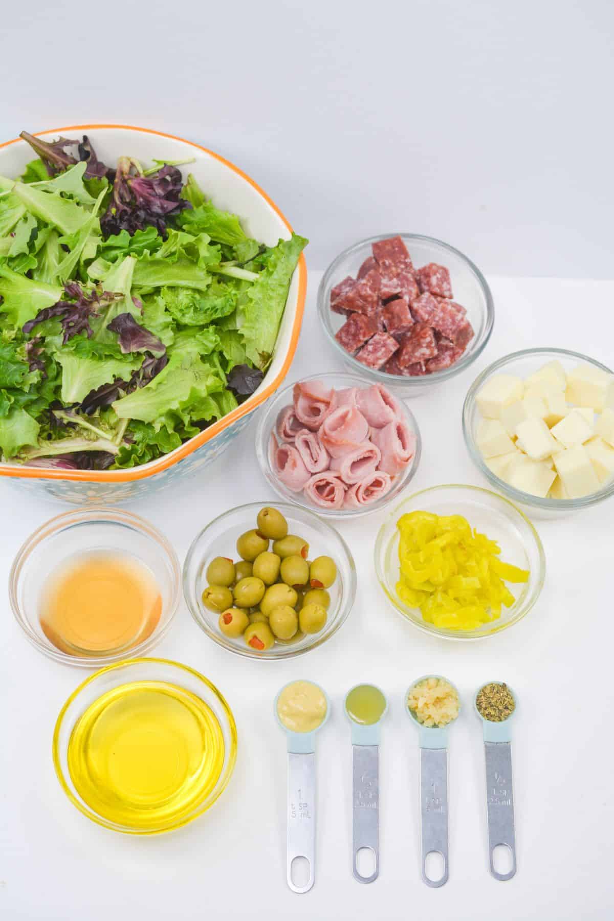 A bowl of mixed greens with small bowls of salami, ham, cheese, olives, and classic Antipasto Misto toppings—pepperoncini, oil, vinegar, mustard, garlic, and Italian seasoning—arranged on a white surface.