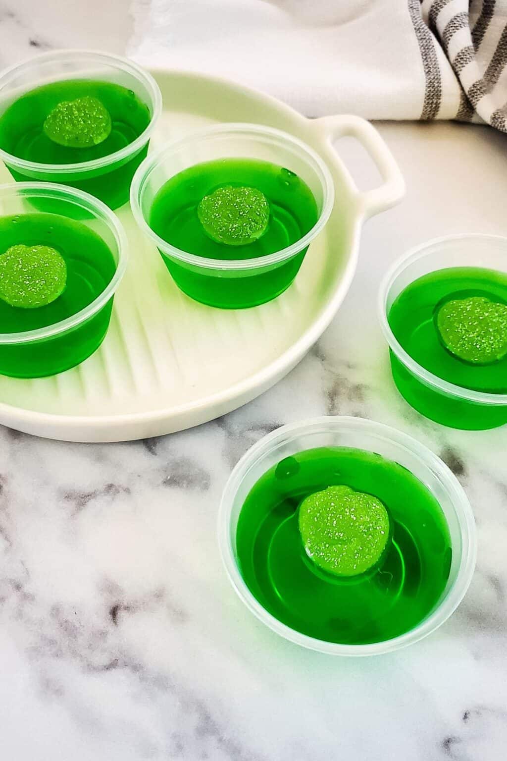 Five small cups of green gelatin, each topped with a green gummy candy, are arranged on a white plate and marble surface with a striped towel in the background—perfectly festive as Halloween Jello Shots for your next spooky celebration.
