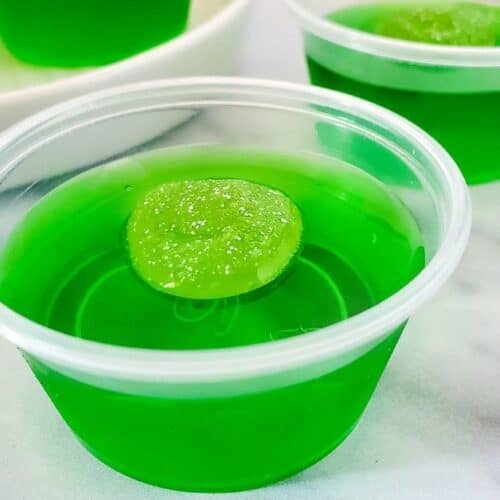A small clear plastic cup filled with bright green gelatin, reminiscent of classic Green Jello Shots, is topped with a green candy and placed on a white surface.
