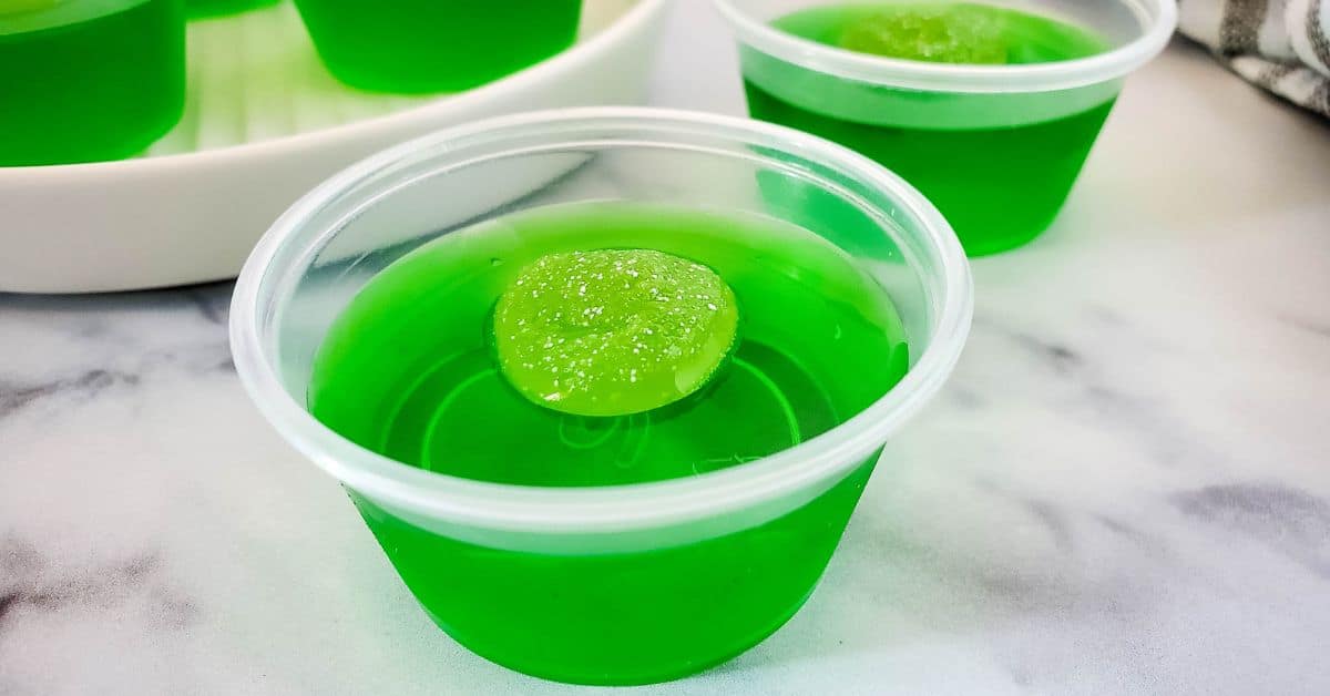 A small clear plastic cup filled with bright green gelatin, reminiscent of classic Green Jello Shots, is topped with a green candy and placed on a white surface.