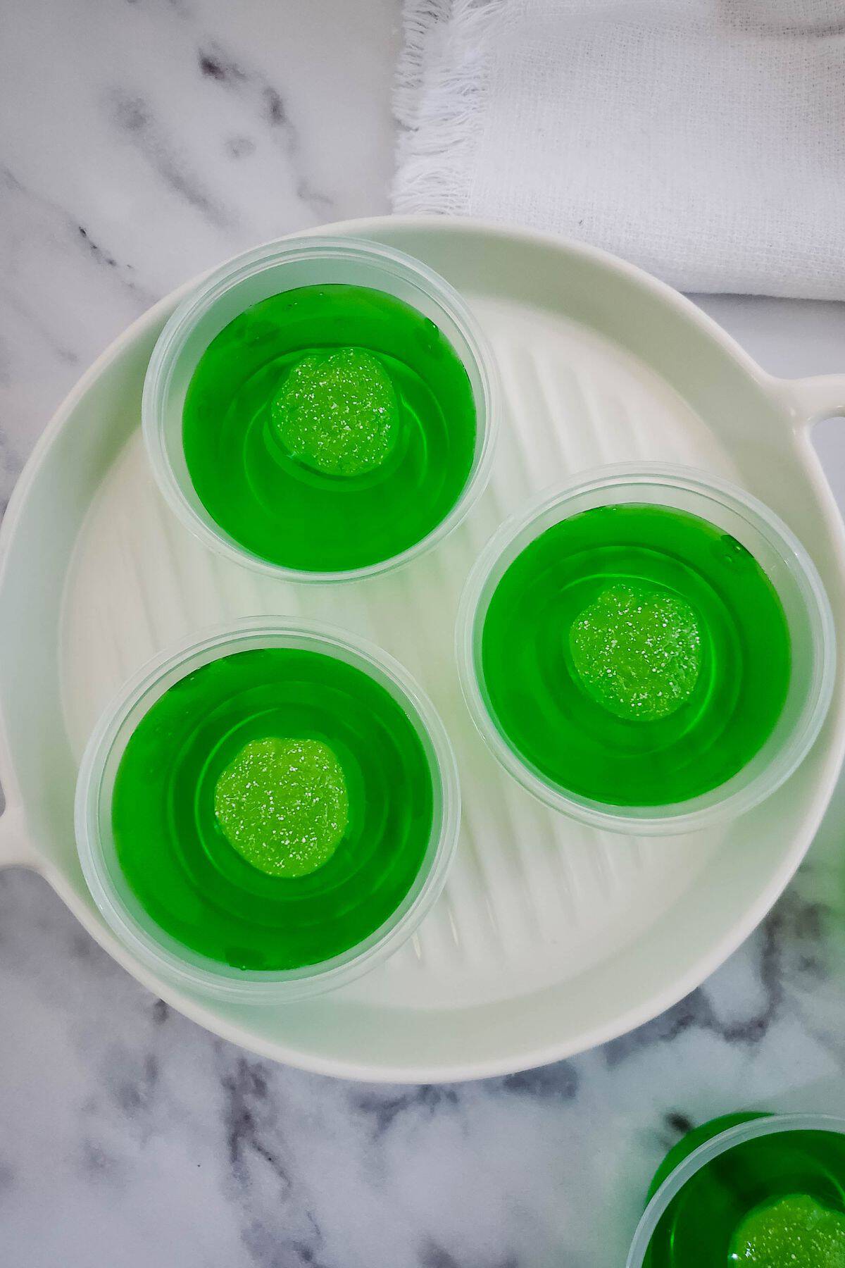 Three plastic cups filled with Green Jello Shots and topped with a green gummy candy, arranged in a white dish on a marble surface.