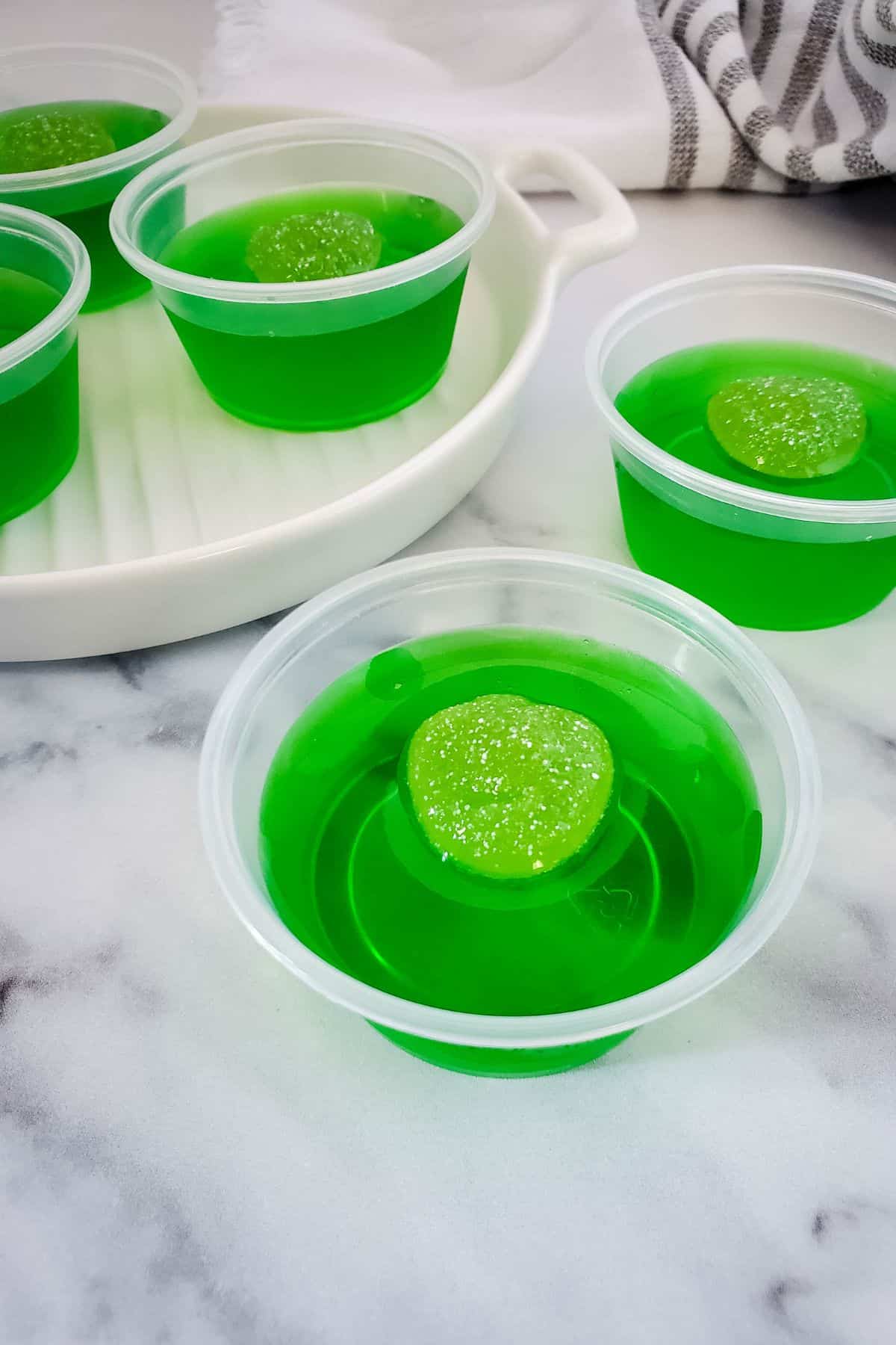 Four plastic cups filled with Green Jello Shots, each topped with a round green candy, placed on a white surface with a striped towel in the background.