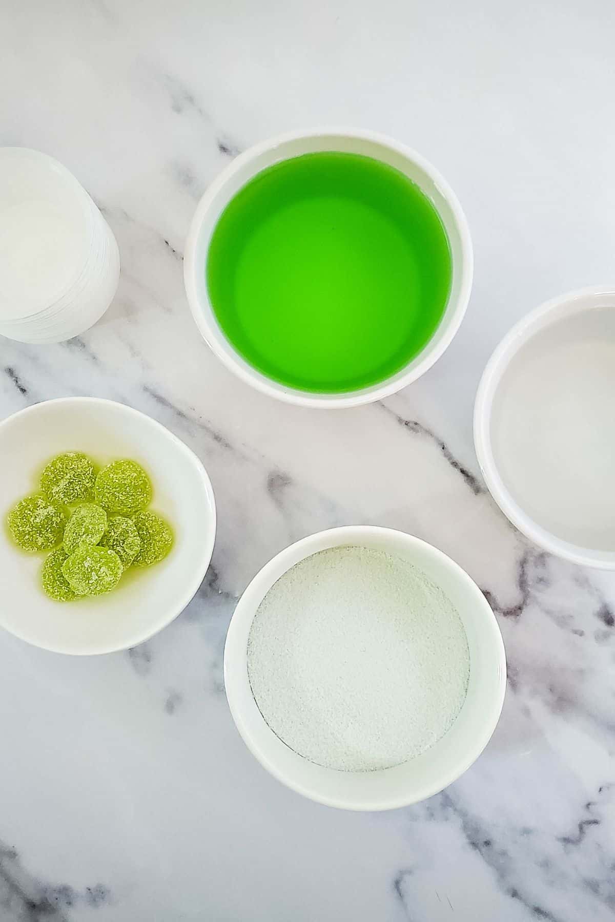Four white bowls on a marble surface, containing green liquid reminiscent of Green Jello Shots, clear liquid, white powder, and green sugared candies. A stack of clear plastic cups is also visible.