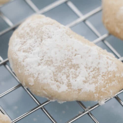 A crescent-shaped Lemon Cooler Cookie covered in powdered sugar rests on a metal cooling rack.