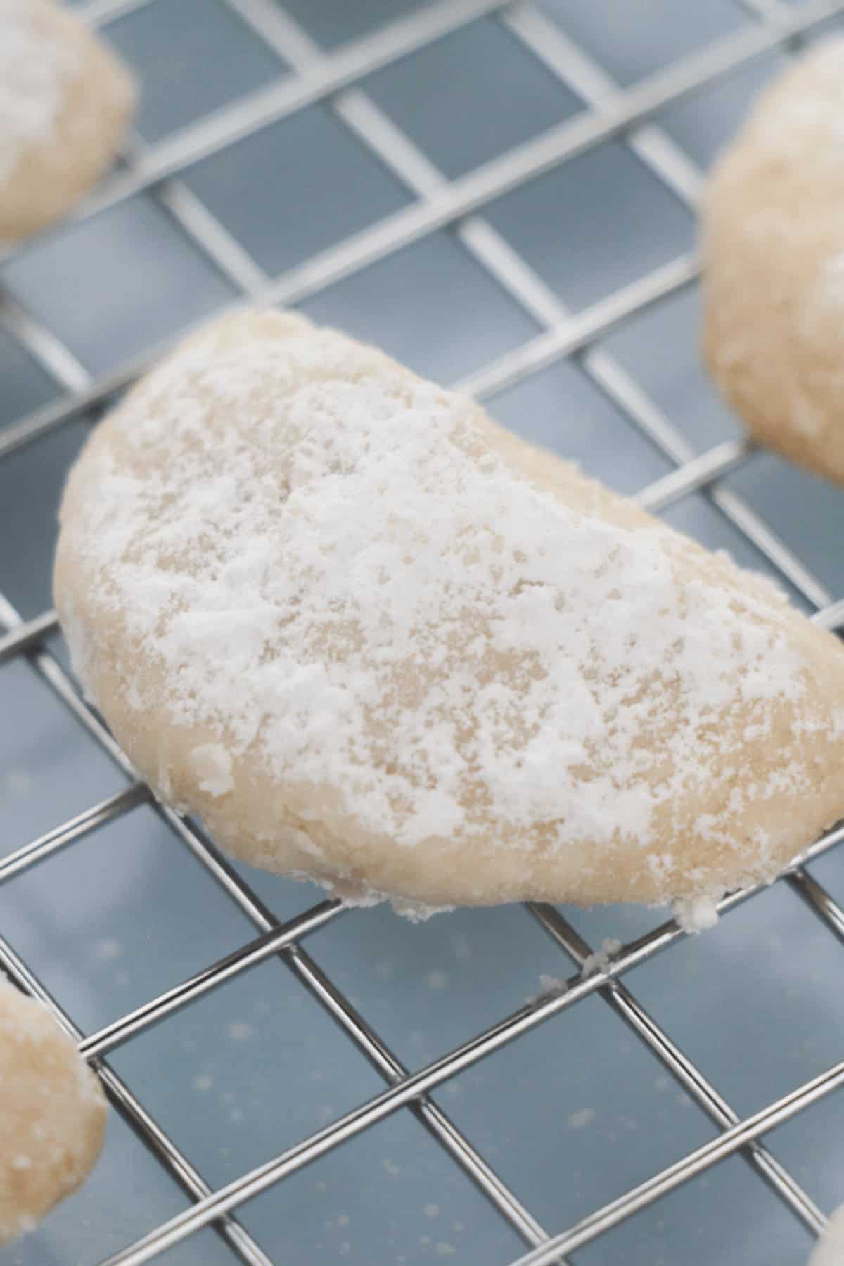 A crescent-shaped Lemon Cooler Cookie covered in powdered sugar rests on a metal cooling rack.