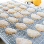 Lemon Cooler Cookies coated in powdered sugar are cooling on a wire rack over a baking sheet, with a lemon and zester visible in the background.