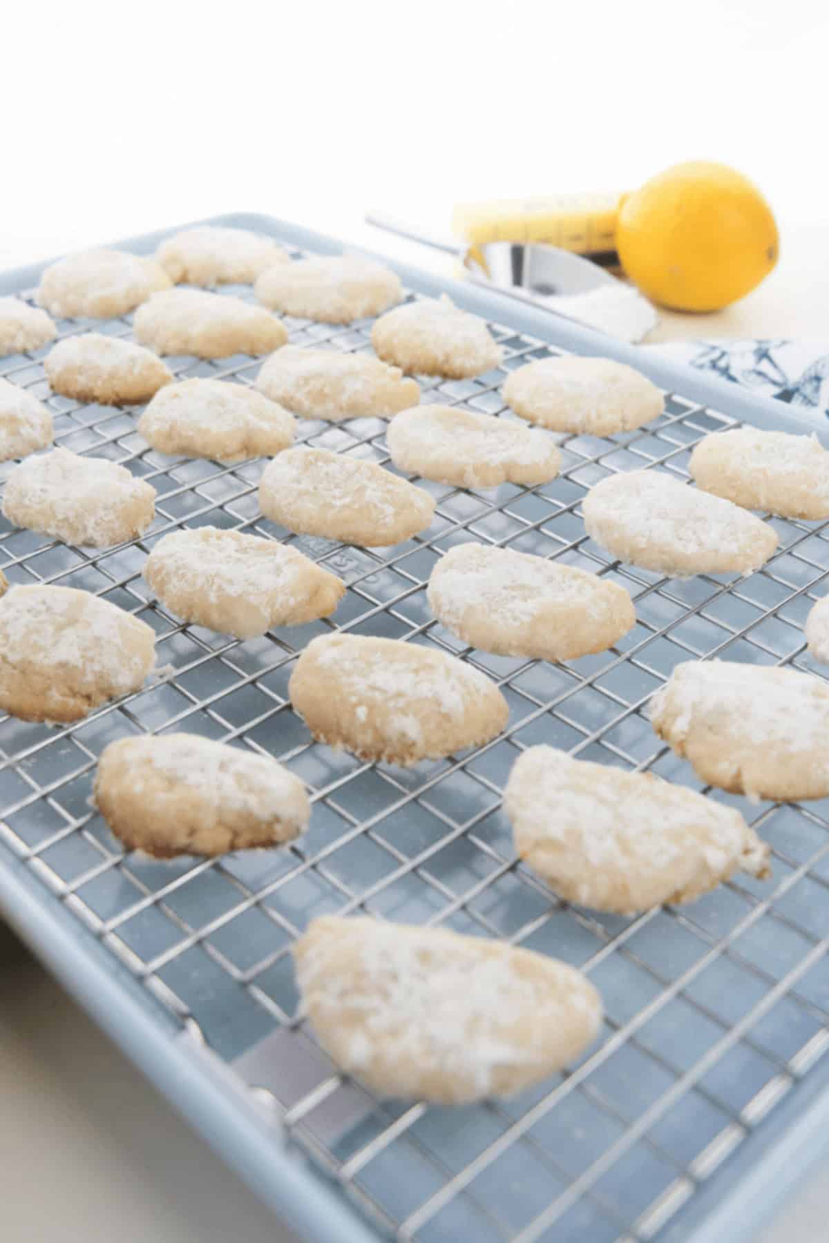 Lemon Cooler Cookies coated in powdered sugar are cooling on a wire rack over a baking sheet, with a lemon and zester visible in the background.