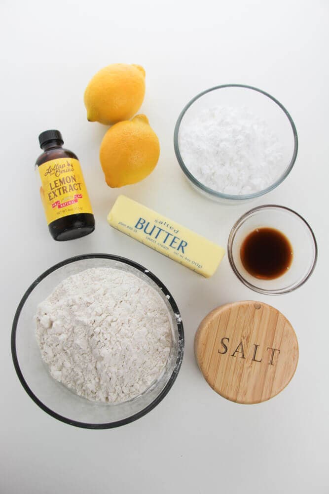 Baking ingredients on a white surface: two lemons, flour, powdered sugar, butter, lemon extract, vanilla extract, and a container of salt.