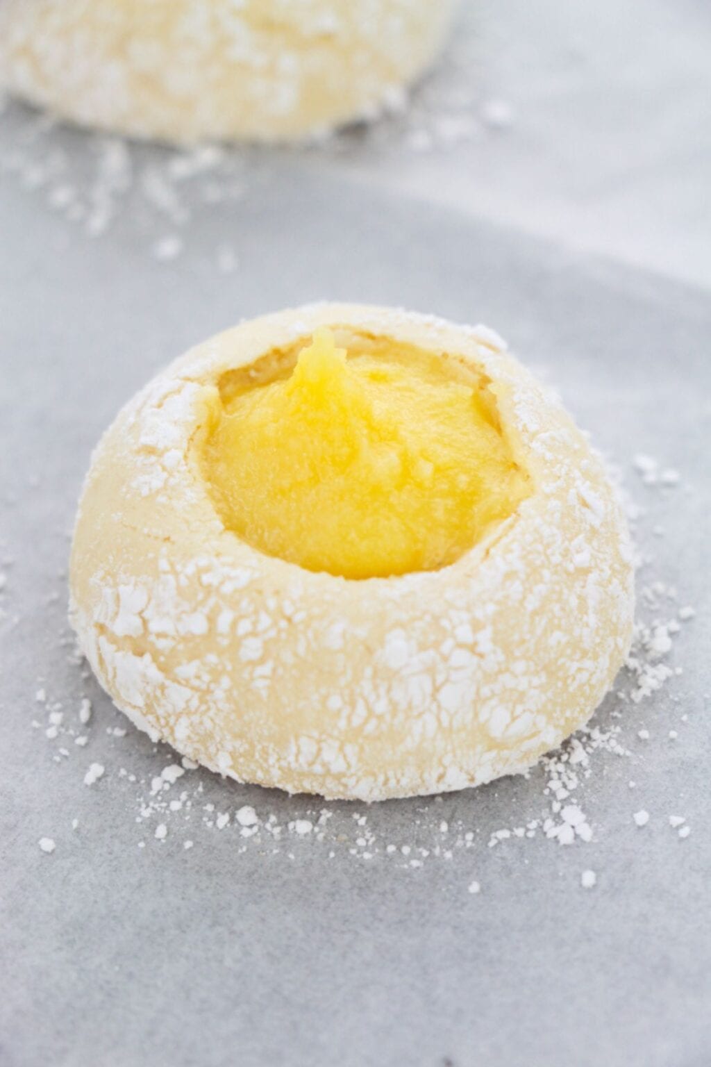 A round, powdered sugar-dusted Lemon Curd Cookie with a yellow custard filling in the center sits on parchment paper.