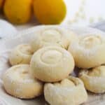 A white plate holds several dusted with powdered sugar, each filled with a creamy center. Whole lemons are visible in the background.