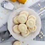 A white plate of Lemon White Chocolate Cookies sits on a gray cloth, surrounded by whole lemons and a scoop of powdered sugar.