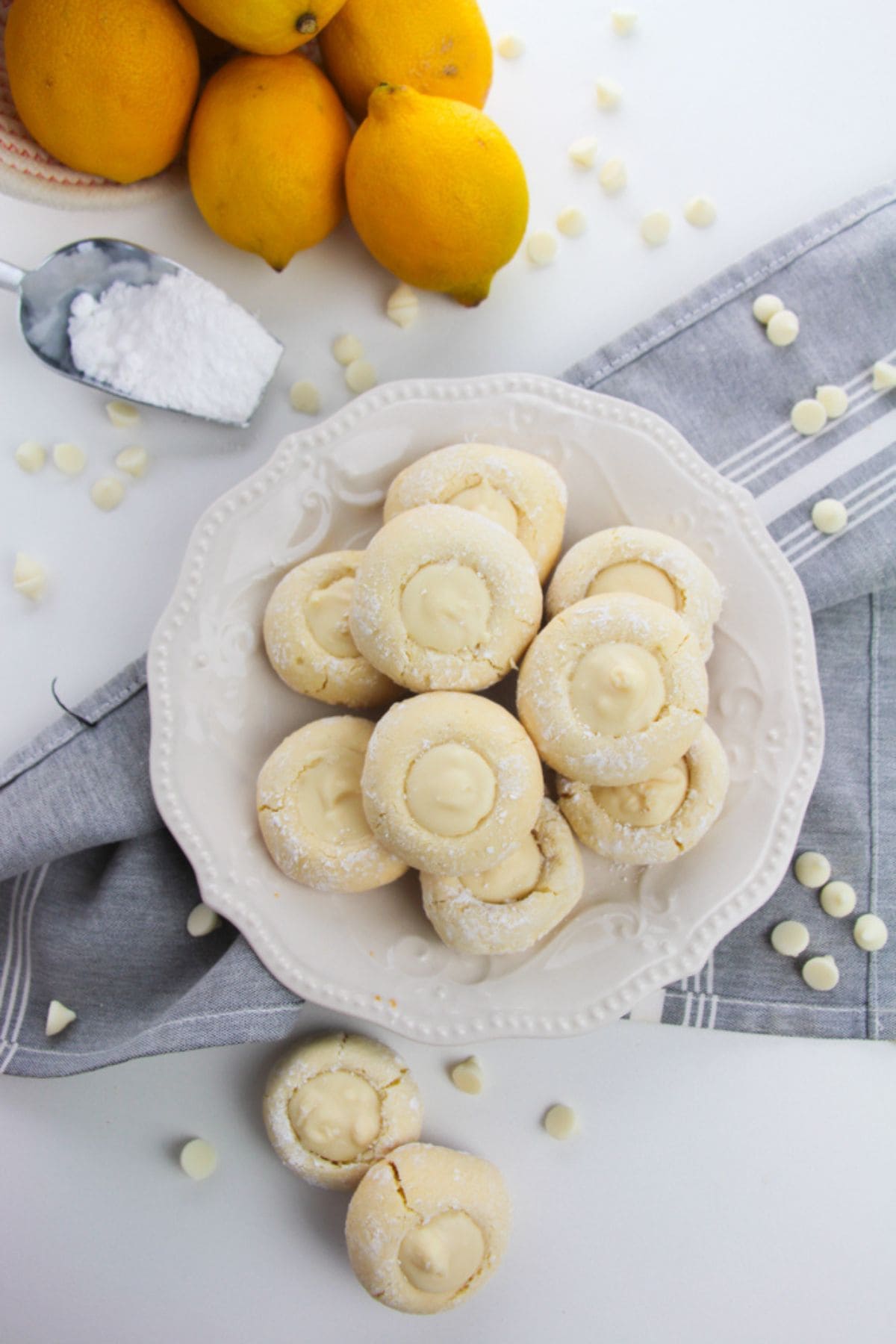 A white plate of Lemon White Chocolate Cookies sits on a gray cloth, surrounded by whole lemons and a scoop of powdered sugar.