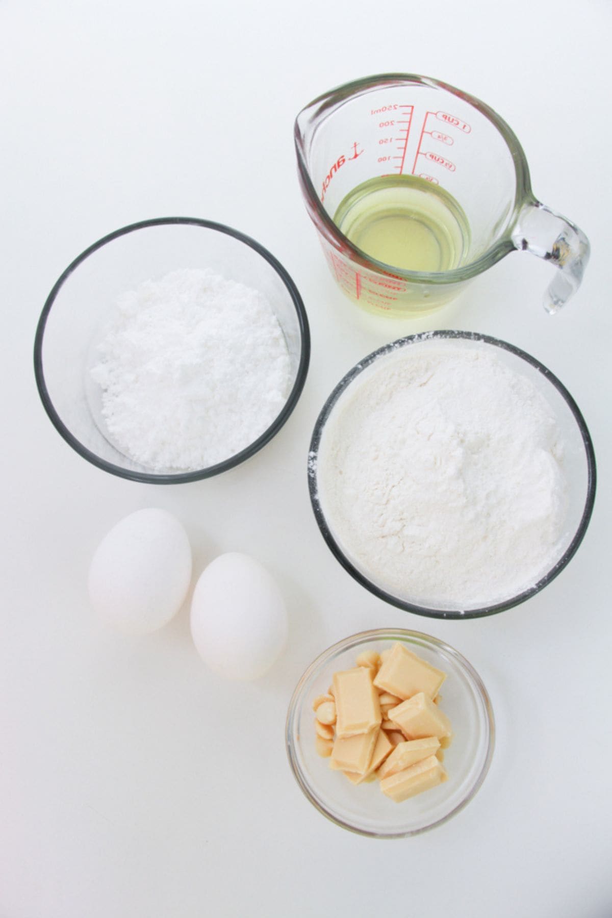 Five baking ingredients for Lemon White Chocolate Cookies on a white surface: two eggs, a bowl of powdered sugar, a bowl of flour, a glass measuring cup with oil, and a bowl of white chocolate pieces.
