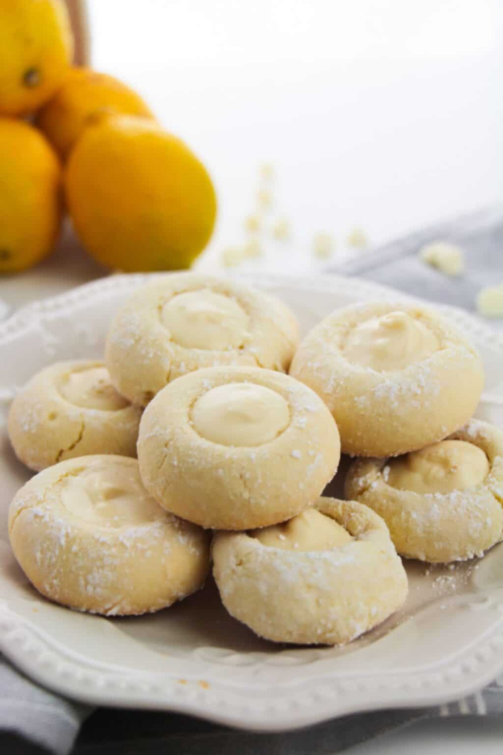 A plate of powdered sugar-dusted Lemon White Chocolate Cookies filled with creamy lemon filling, with whole lemons in the background.