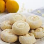 A plate of powdered sugar-dusted Lemon White Chocolate Cookies filled with creamy lemon filling, with whole lemons in the background.